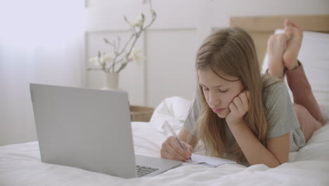Kleines-Mädchen-Zeichnet-Und-Schreibt-In-Einem-Heft,-Liegt-Auf-Dem-Bett-Vor-Dem-Laptop-In-Ihrem-Zimmer-Und-Wartet-Auf-Den-Online-Unterricht